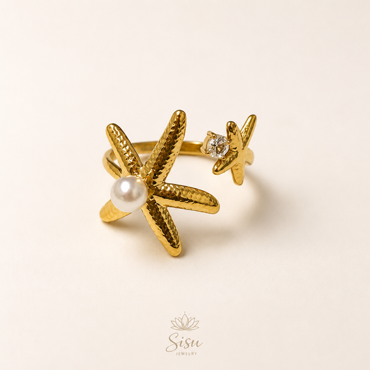 Anillo dorado con estrella de mar, perla y detalle brillante sobre fondo crema elegante de Sisu Jewelry.