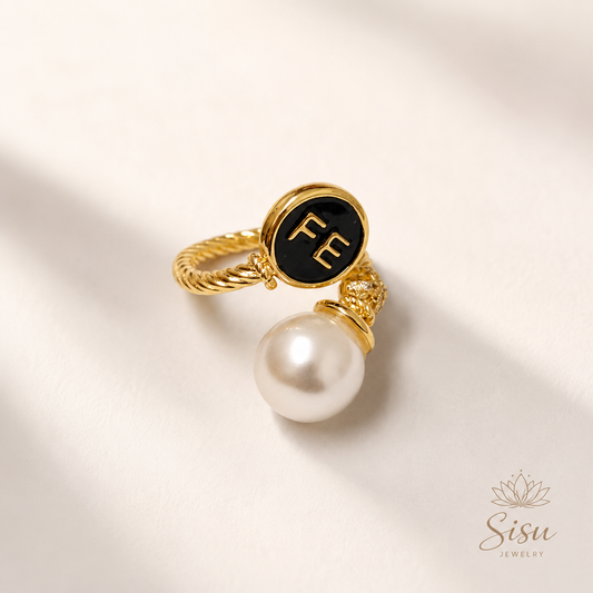 Anillo dorado con detalle FE y perla blanca sobre fondo crema elegante de Sisu Jewelry.